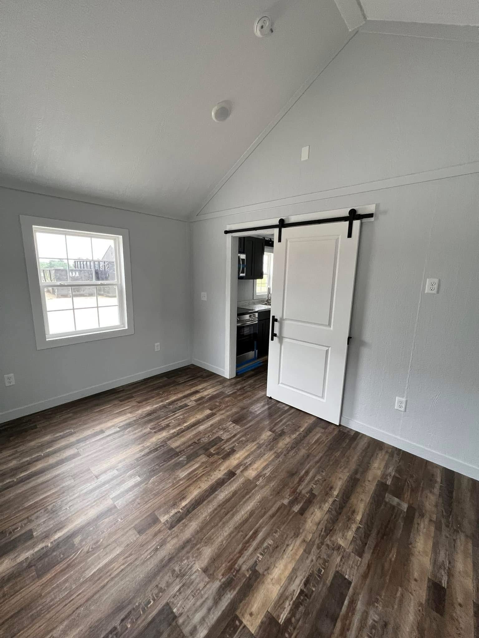 Empty room with light gray walls, wooden flooring, a large window on the left, and a sliding white barn door leading to a small kitchen area. Minimalist and clean atmosphere.