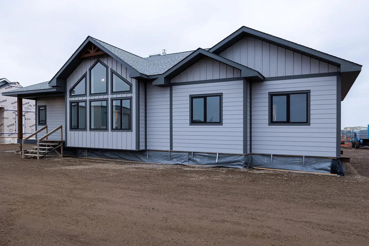 Single-story gray house with modern, large triangular windows and black trim. It has a porch with steps, set on bare dirt under an overcast sky.