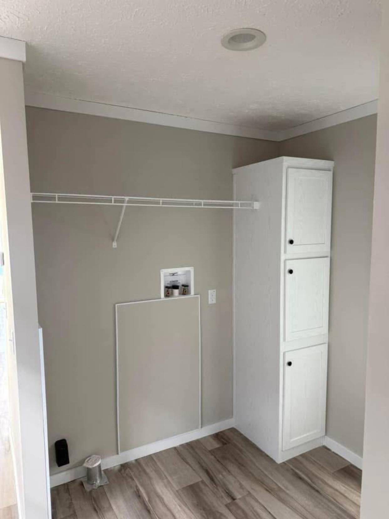 Laundry room with beige walls and wood flooring, featuring white cabinets with black handles and an empty wire shelf above a utility hookup.