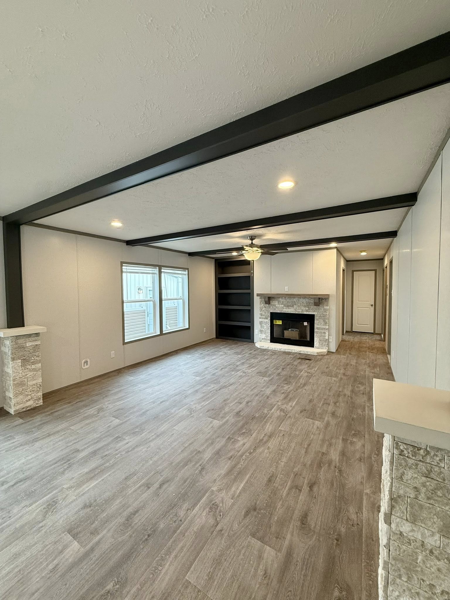 Spacious living room with light wood flooring, a stone fireplace, built-in shelves, and a ceiling fan. Large windows illuminate the neutral space.