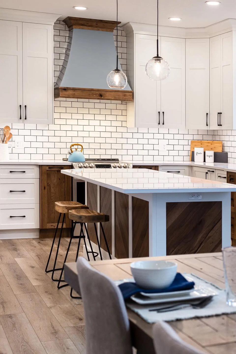 Modern kitchen with white cabinets, subway tile backsplash, and wood accents. Island with bar stools, pendant lights, and place settings on table.