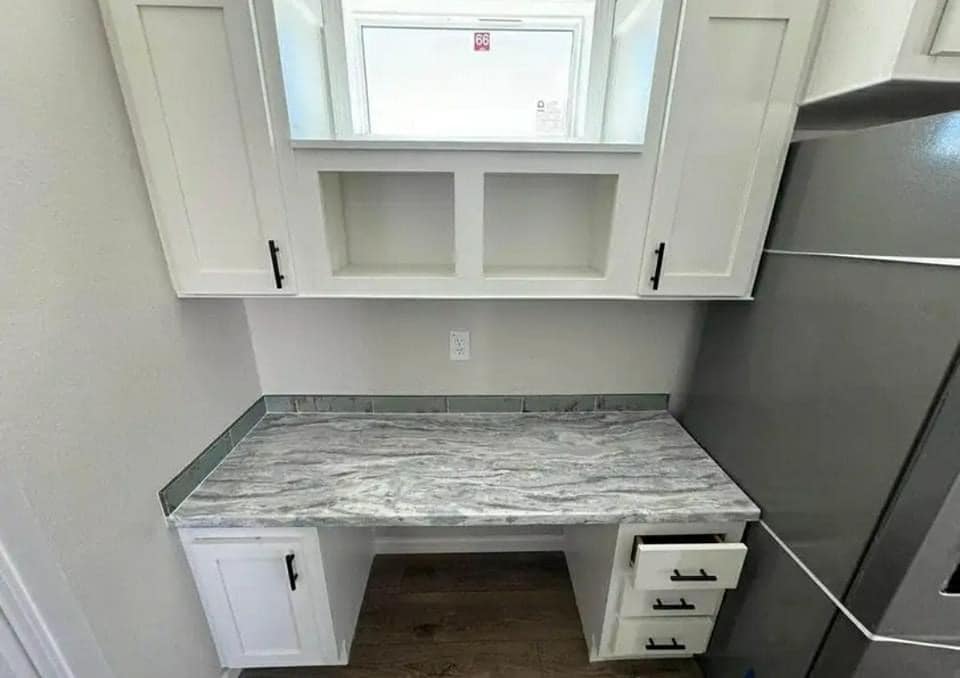 Compact desk space with marble countertop, set between white cabinetry. Features built-in shelves above and drawers to the right. Bright and organized.