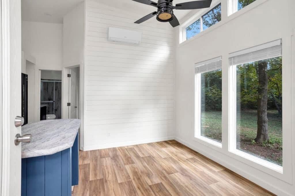 Modern room with high ceilings, large windows overlooking a garden, wood flooring, a blue kitchen counter, white walls, and a black ceiling fan.