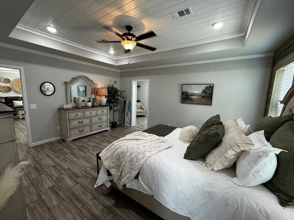 Cozy bedroom with a tray ceiling and fan, featuring a neatly made bed with green and white pillows. A dresser with a mirror and lamp is on the left.