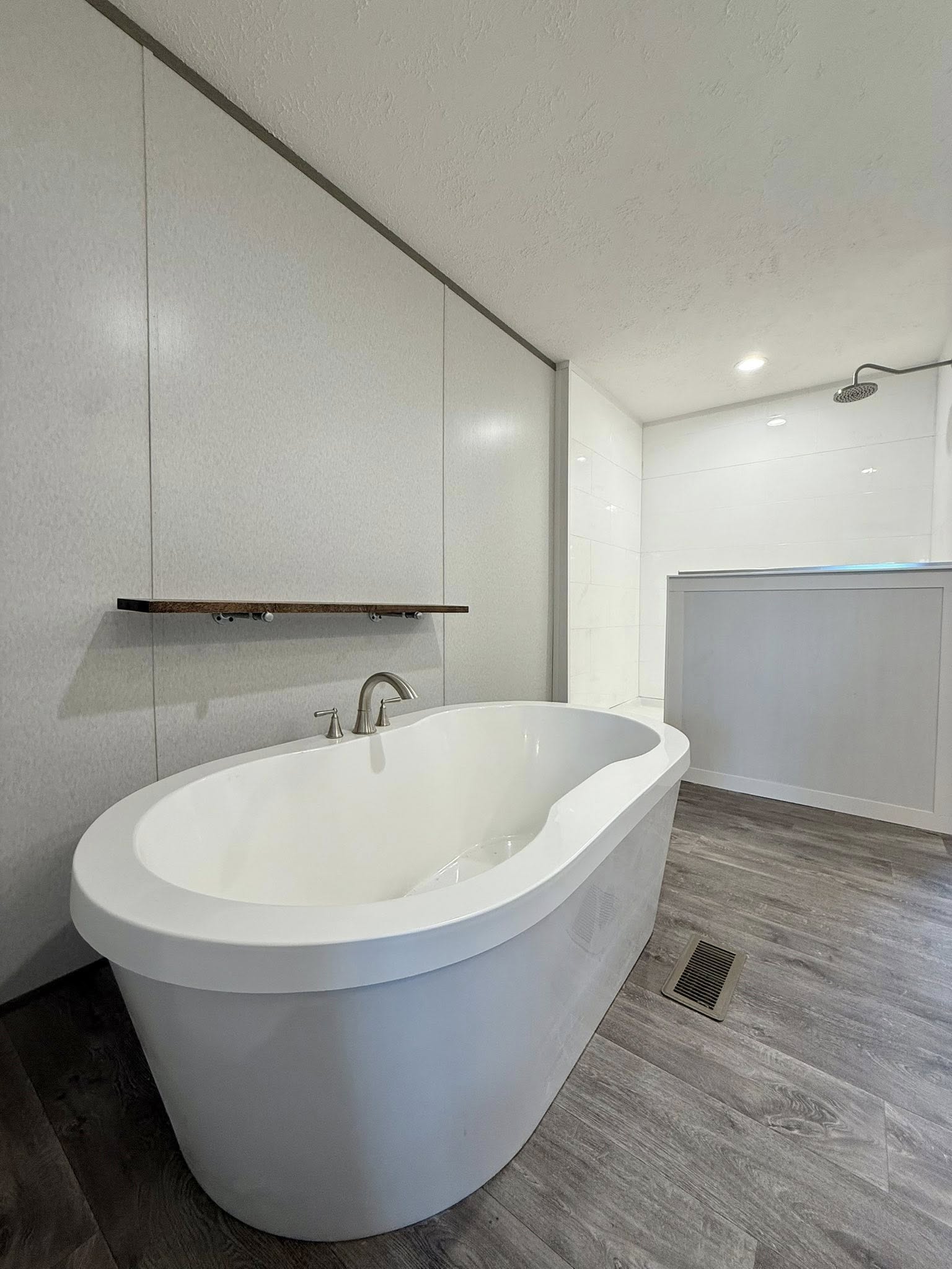 Modern bathroom featuring a sleek white freestanding bathtub on dark wood flooring. Overhead, spotlights illuminate a serene shower area.