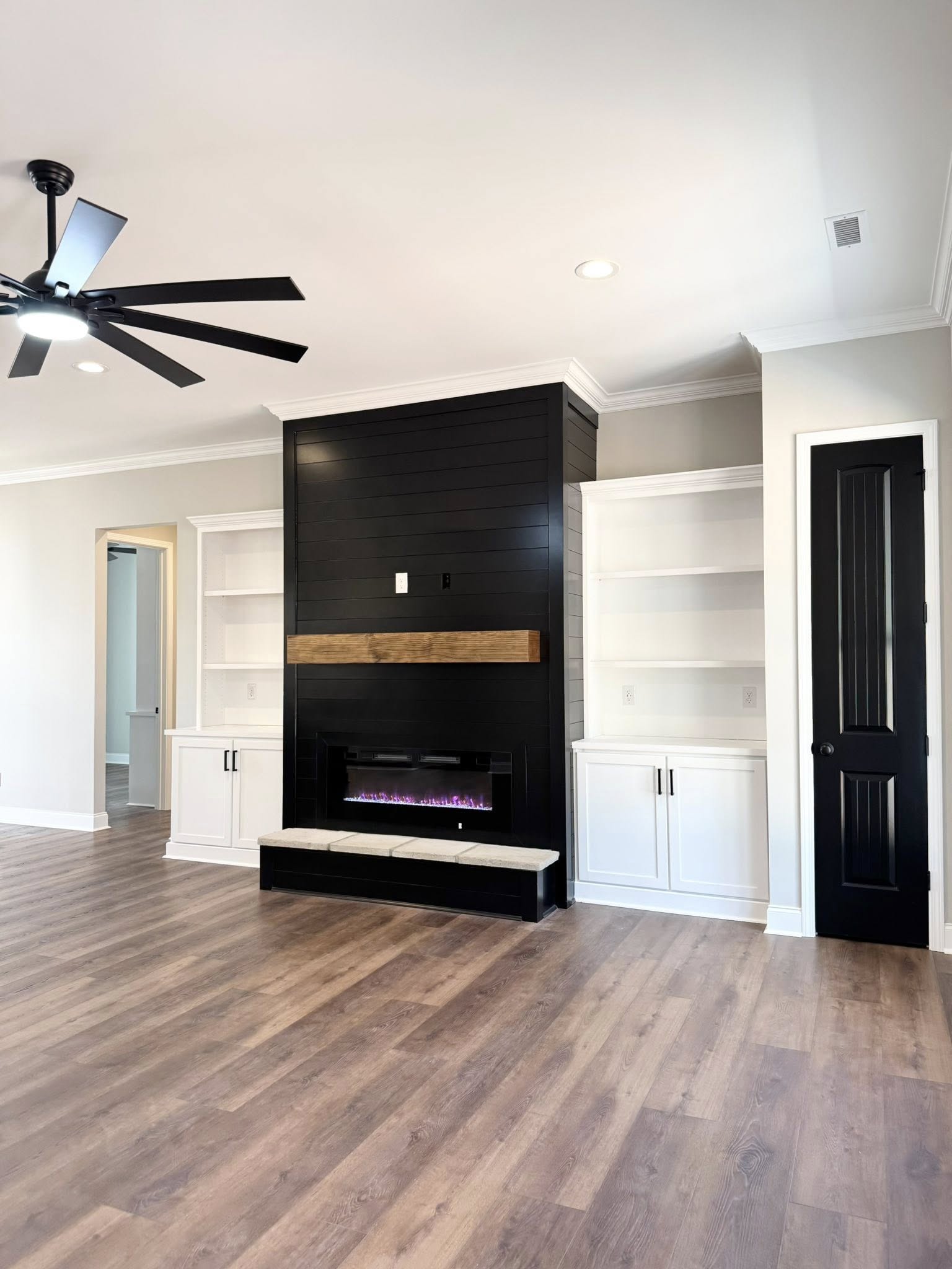 Spacious living room with wood flooring, large ceiling fan, modern black fireplace with wooden mantel, open shelving, and sliding glass doors. Bright and minimalistic.