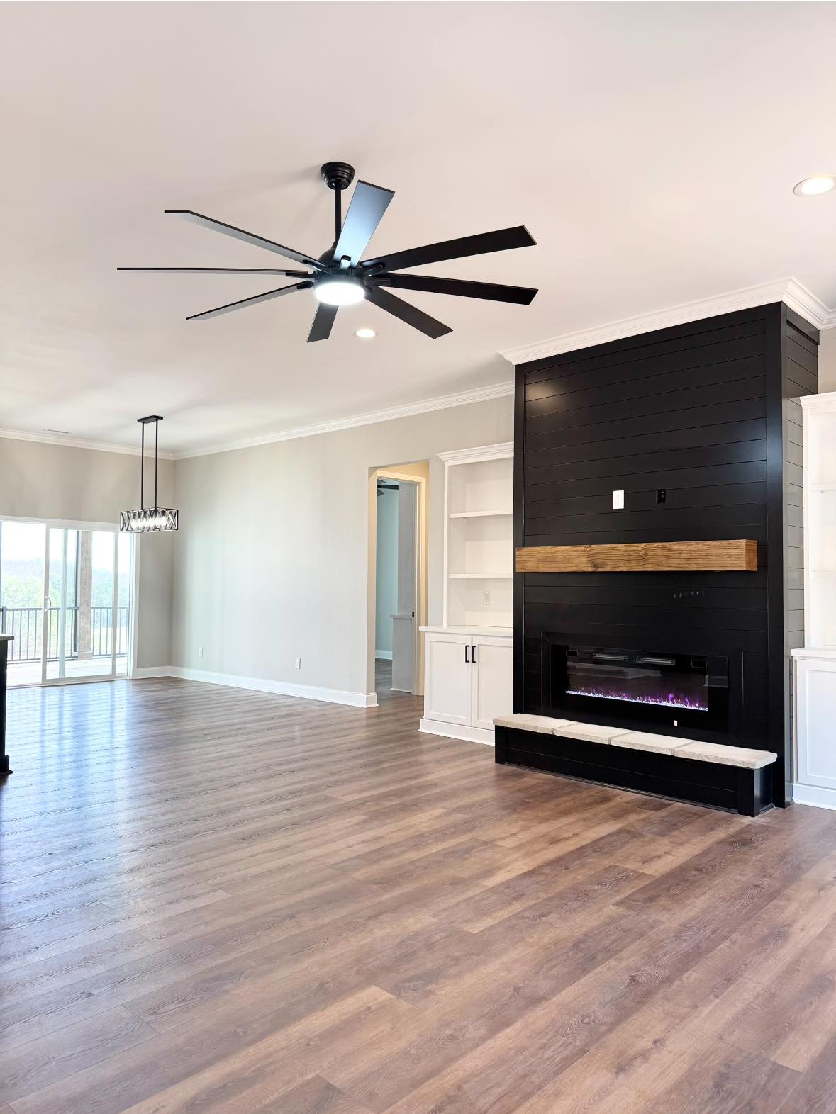 Spacious modern living room with wood floors, large ceiling fan, black shiplap fireplace, open shelves, and glass doors leading to a balcony. Bright and airy.