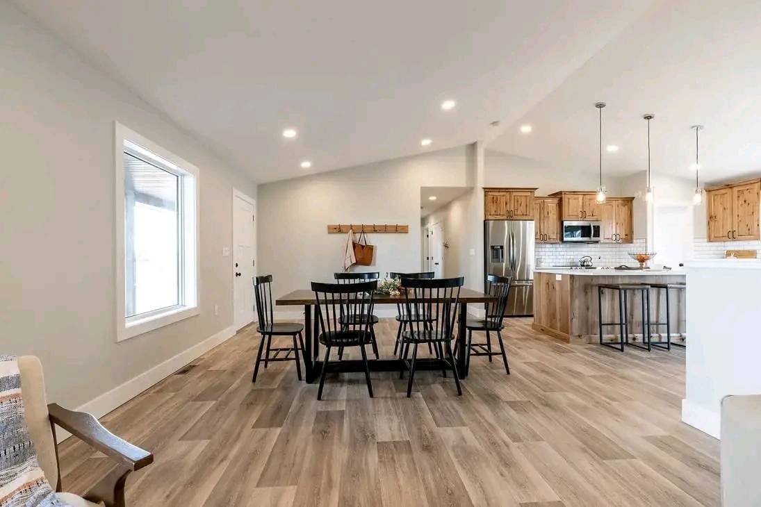 Spacious open-plan dining and kitchen area with wooden floors. Features a large wooden table, black chairs, hanging pendant lights, and rustic cabinets. Cozy and inviting atmosphere.