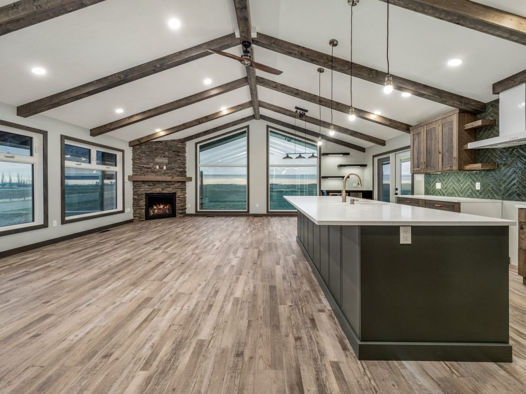Spacious modern kitchen with vaulted ceiling, exposed wooden beams, and large windows. Features a stone fireplace, long island, and pendant lights. Cozy ambiance.