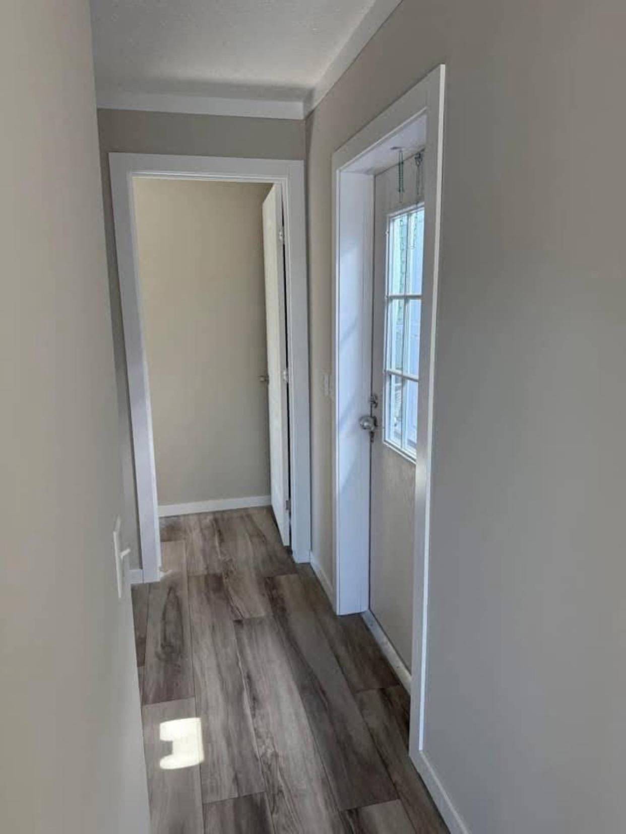 Narrow hallway with light gray walls and wood flooring, leading to a doorway. Sunlight streams in through a glass door on the right, creating a cozy ambiance.