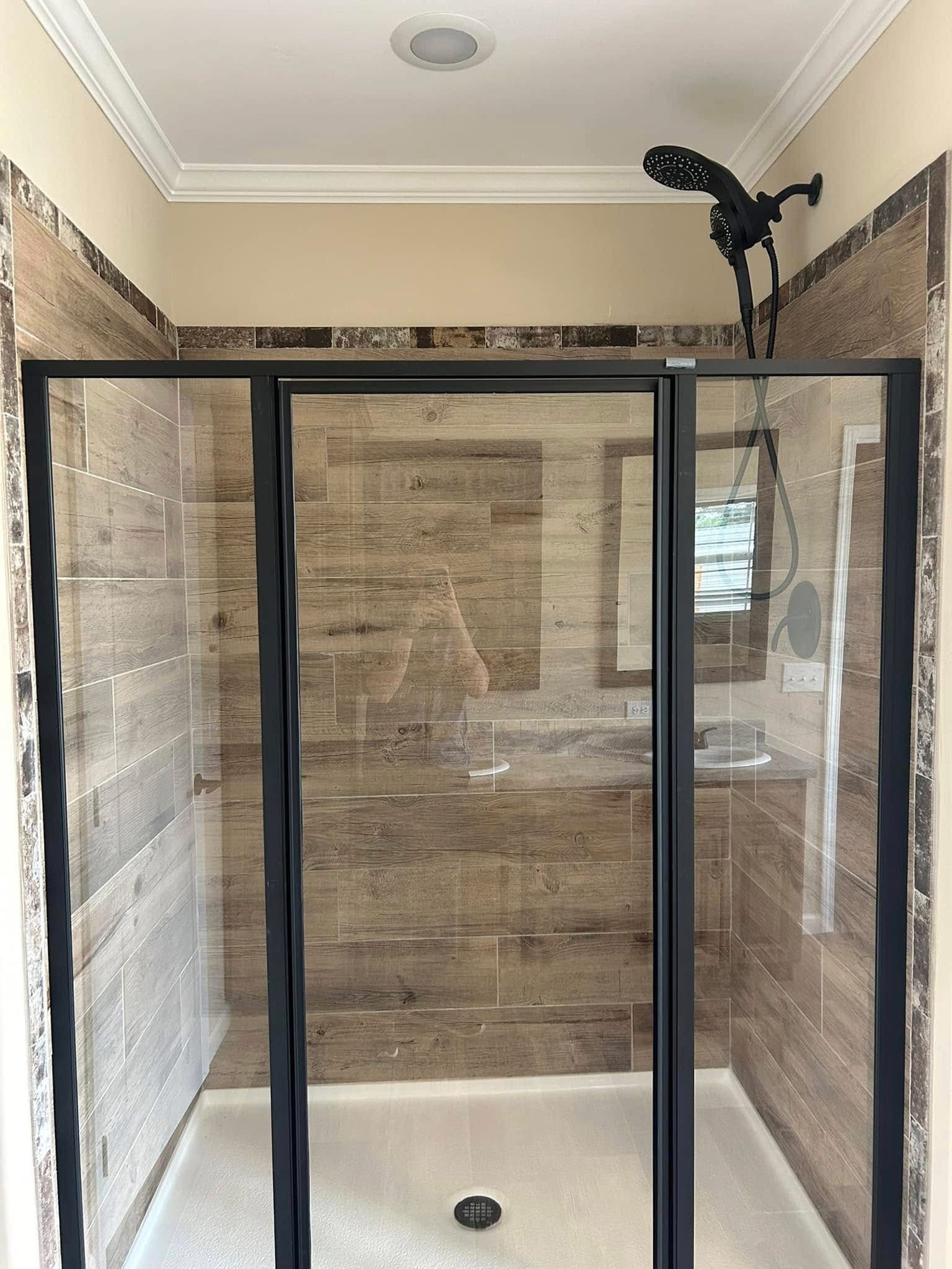 Modern shower with dark-framed glass doors and wood-look tiles. Overhead black showerhead. Neutral tones and a tidy, inviting ambiance.