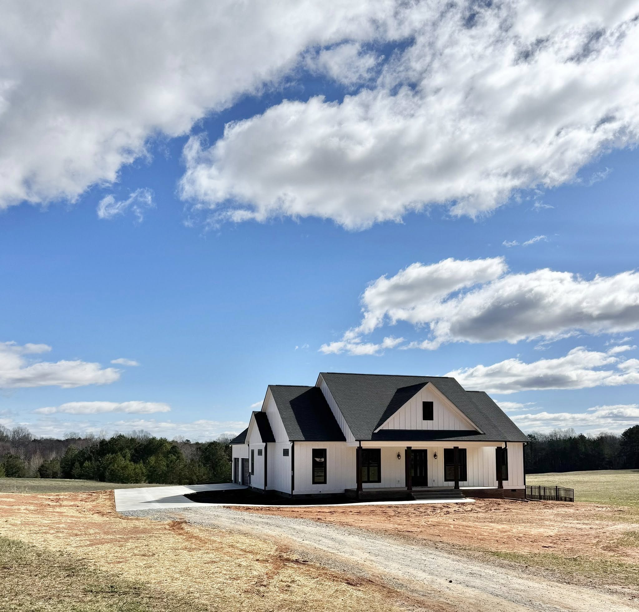 A modern white house with a dark roof stands in an open field under a bright blue sky with scattered clouds, conveying a serene, rural setting.