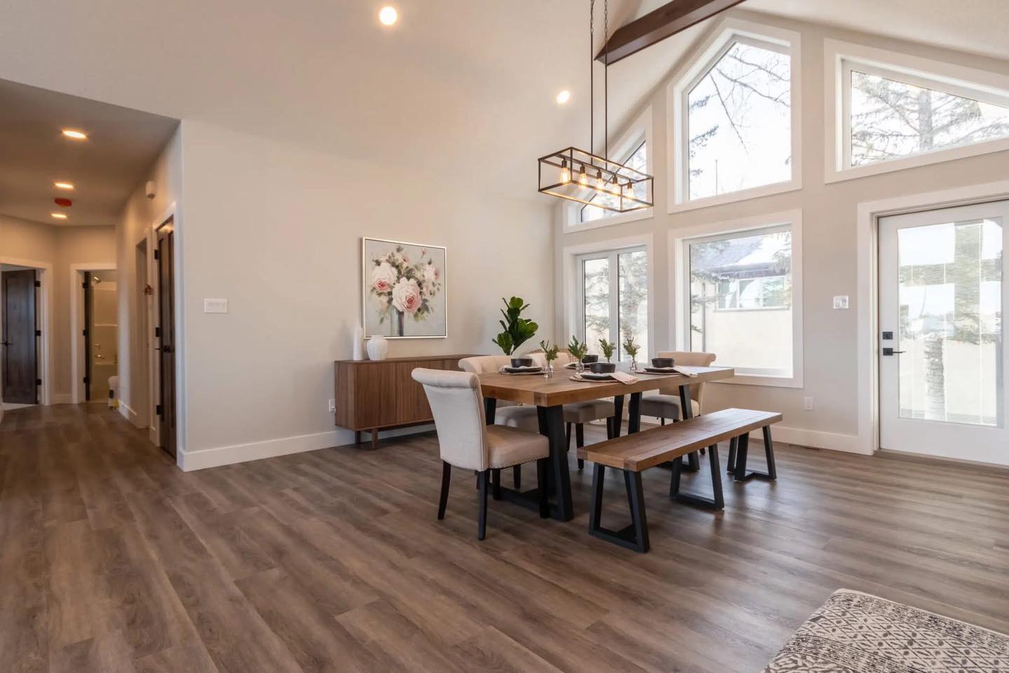 Modern dining room with vaulted ceilings and large windows, featuring a wooden table, benches, upholstered chairs, and a floral painting, evoking a cozy, elegant ambiance.