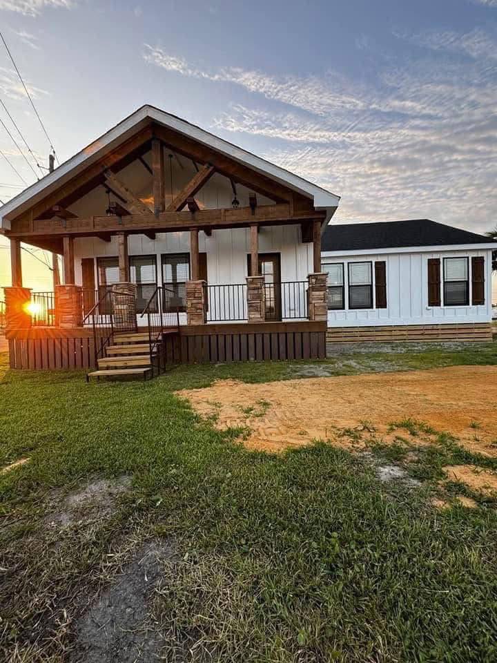 A charming white house with a wooden porch at sunset. Warm light creates a cozy atmosphere. Green grass and a patch of bare earth in front.