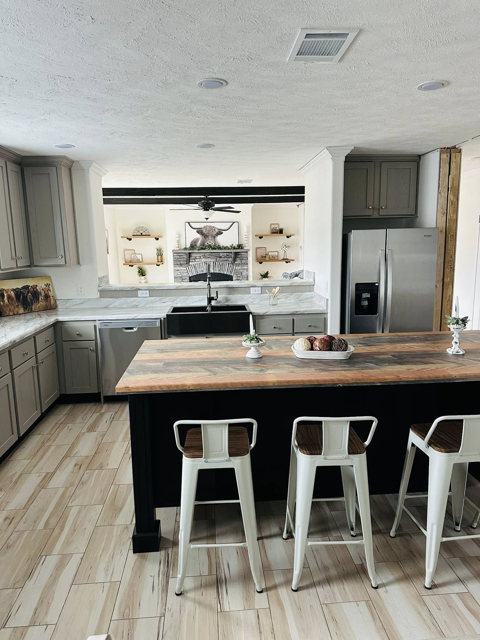 Modern kitchen with a central island featuring a wooden countertop and three white stools. Gray cabinets, stainless steel appliances, and a farmhouse sink accentuate the space. Shelves with decor, including plants and a bull sculpture, are visible in the background, creating a welcoming, rustic atmosphere.