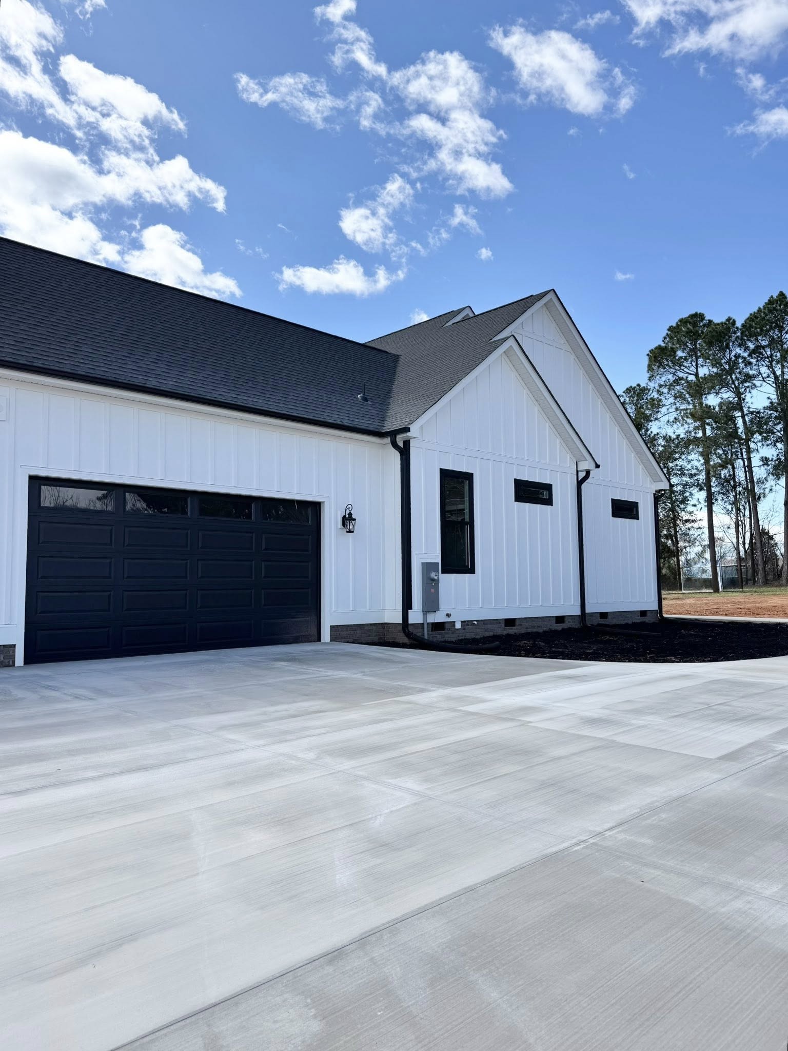 Modern white house with black garage door, gable roof, and concrete driveway. Clear blue sky with clouds and pine trees in the background.