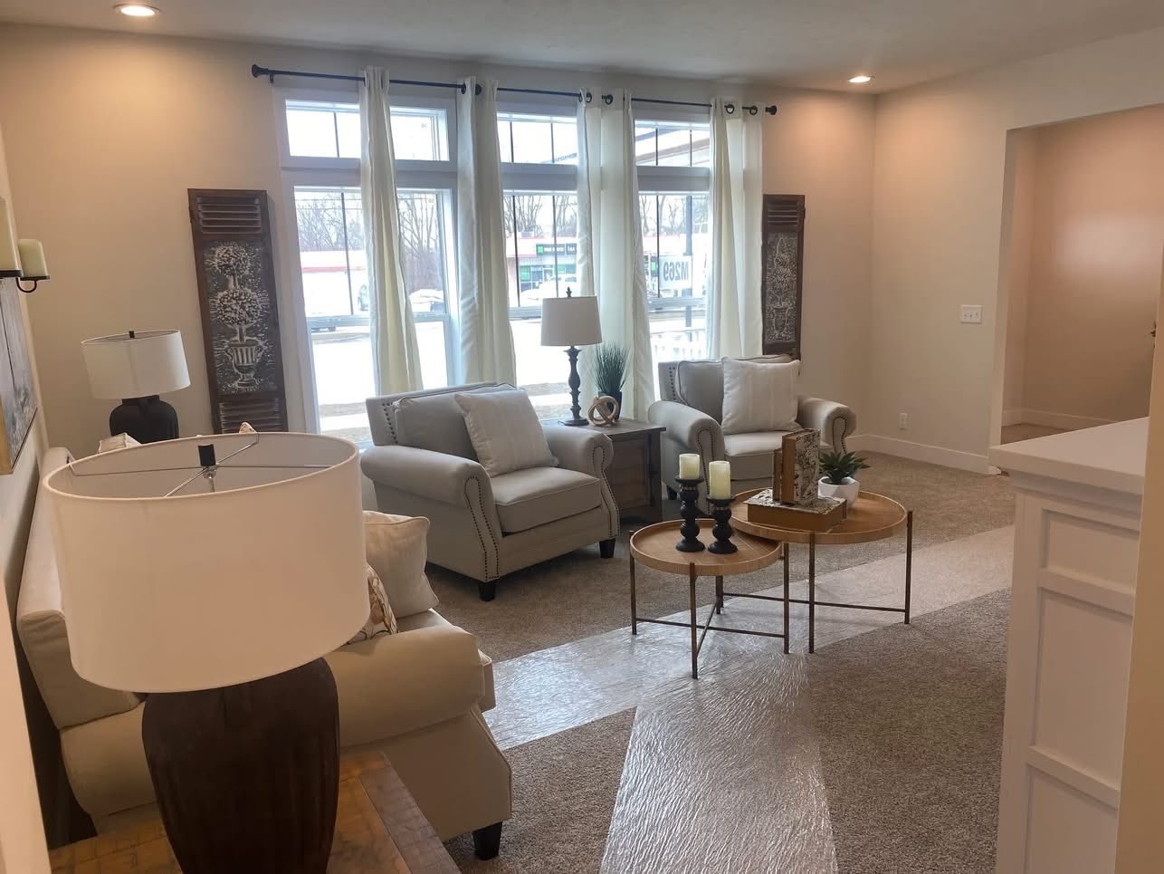 Cozy living room with cream sofas, large windows with curtains, and two round wooden tables. Soft lighting creates a warm, inviting atmosphere.
