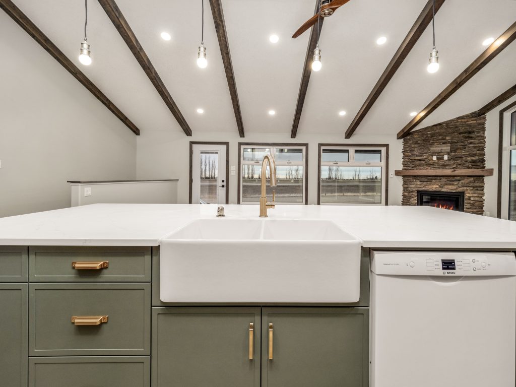 Modern kitchen with a large white island, farmhouse sink, and gold faucet. Exposed beams, pendant lights, and a stone fireplace create a cozy ambiance.