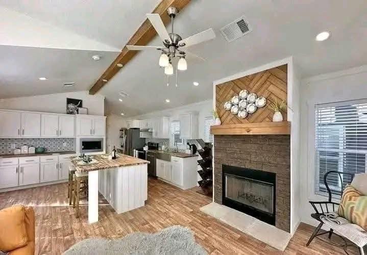 Spacious, cozy kitchen with white cabinets, wood flooring, island with bar stools, and stone fireplace. Modern, warm, inviting atmosphere.