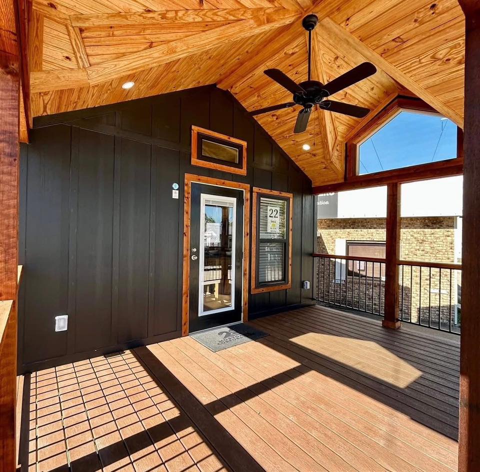 Covered porch with a wood ceiling and floor, black walls, and glass door. Ceiling fan hangs above, creating a cozy, inviting outdoor space.