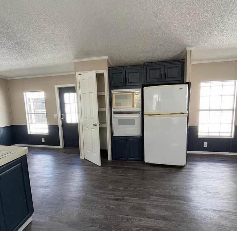 A spacious kitchen with two large windows, dark wood flooring, a white fridge, and an integrated oven. Cabinets are dark blue, and a pantry door is open.
