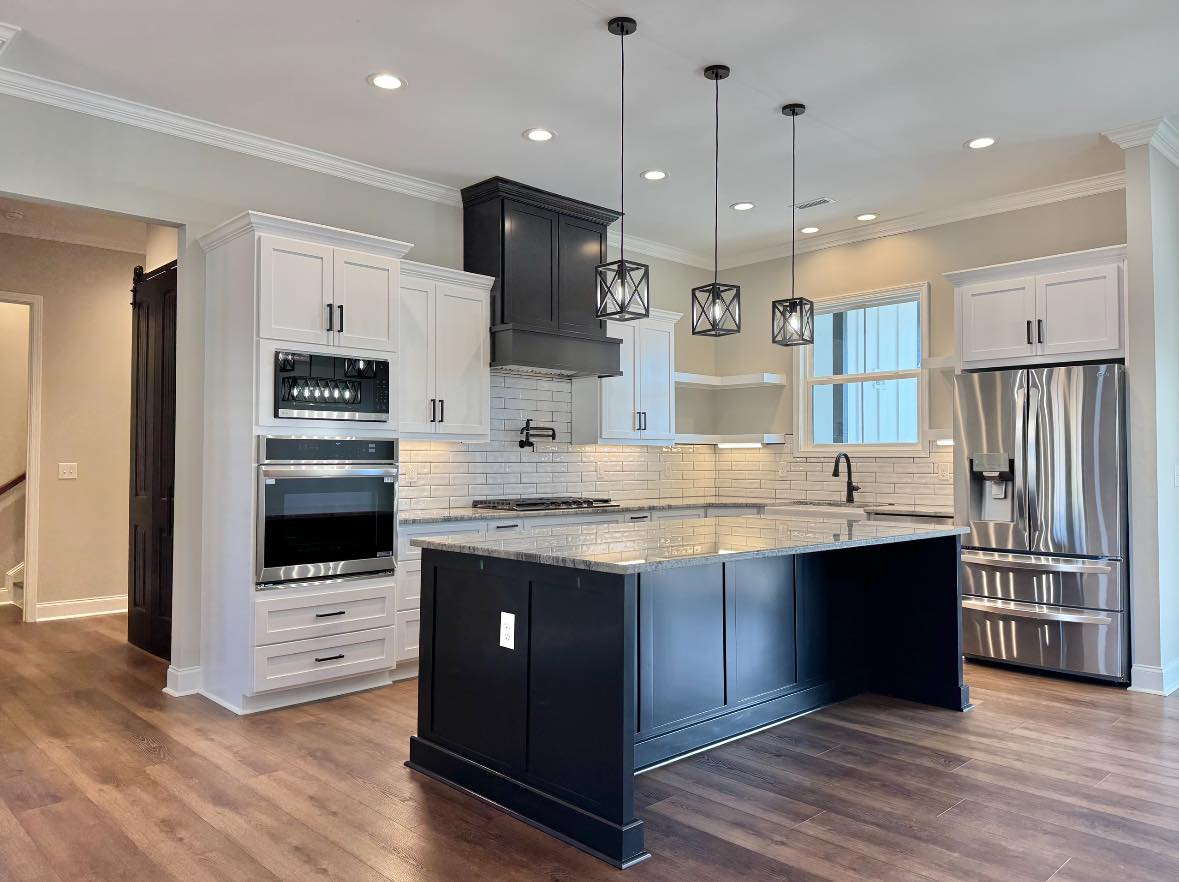 Modern kitchen with dark hardwood floors, white cabinets, and a large black island. Features stainless steel appliances and pendant lights. Bright and sleek.