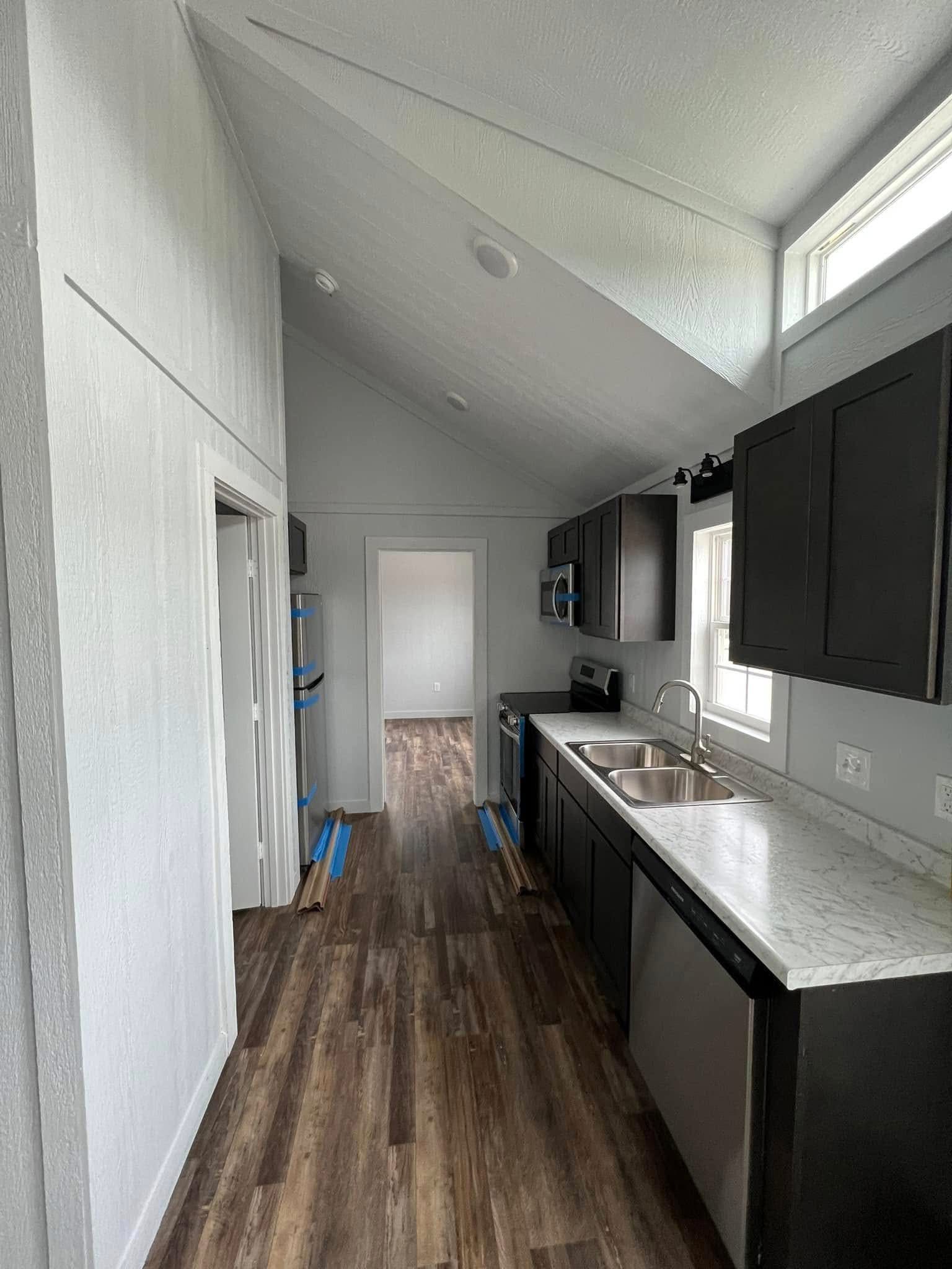Modern kitchen interior with a vaulted ceiling, dark cabinets, light marble countertops, and wood flooring. Natural light streams through a window.