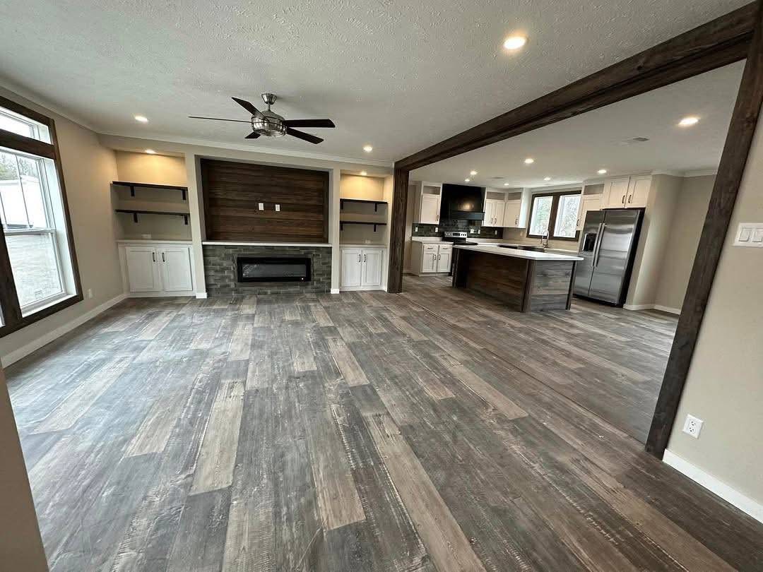 Spacious open-plan living area with wood-style flooring, modern kitchen, and built-in fireplace. Warm lighting creates a cozy, inviting atmosphere.