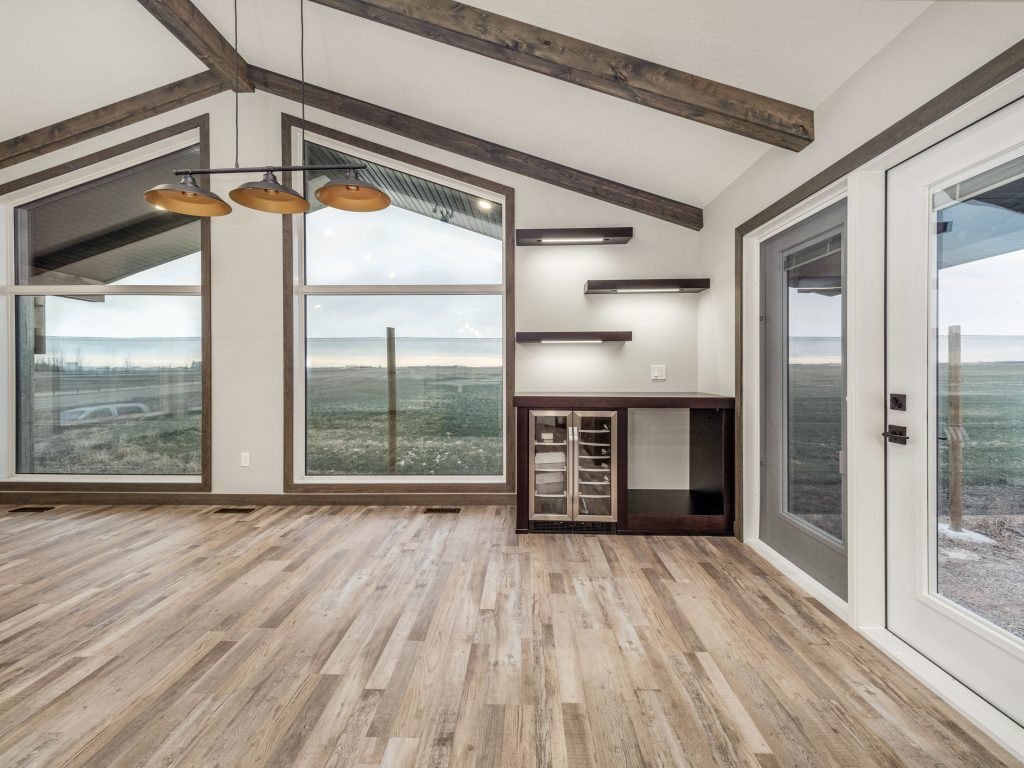 Spacious room with large windows, offering a scenic outdoor view. Features wood floors, ceiling beams, modern lighting, and sleek built-in shelves. Calm ambiance.