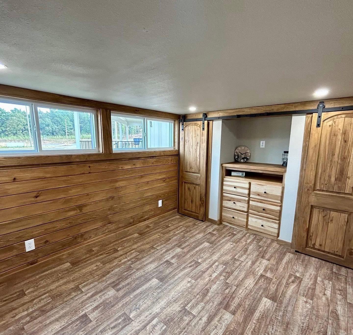 Spacious room with wooden walls and floor, large windows on the left, built-in wooden drawers, and sliding barn doors, creating a rustic vibe.