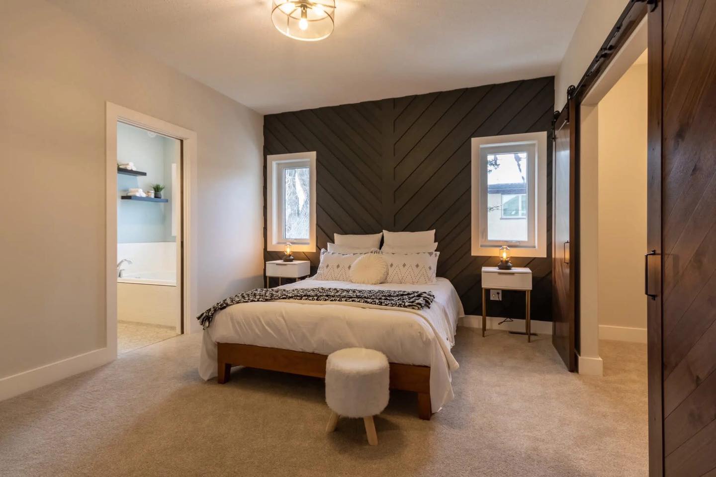 Cozy bedroom with a modern aesthetic. Features a white bed with decorative pillows, dark accent wall, large windows, soft lighting, and a fluffy stool. Warm, inviting vibe.