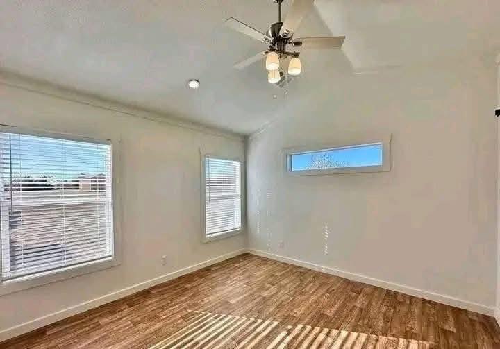 Bright room with wooden floors, white walls, and three windows allowing natural light. Ceiling fan with lights hangs above, creating a cozy, airy feel.