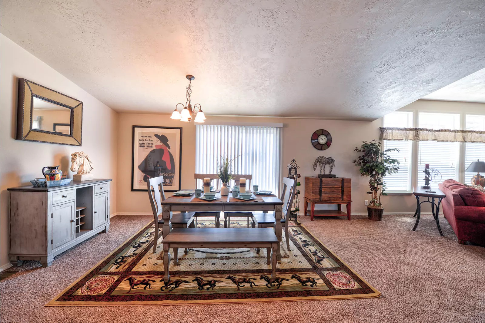 Dining room with a rustic table set for four on a patterned rug. Soft lighting, wall art, and decor create a warm, inviting atmosphere.