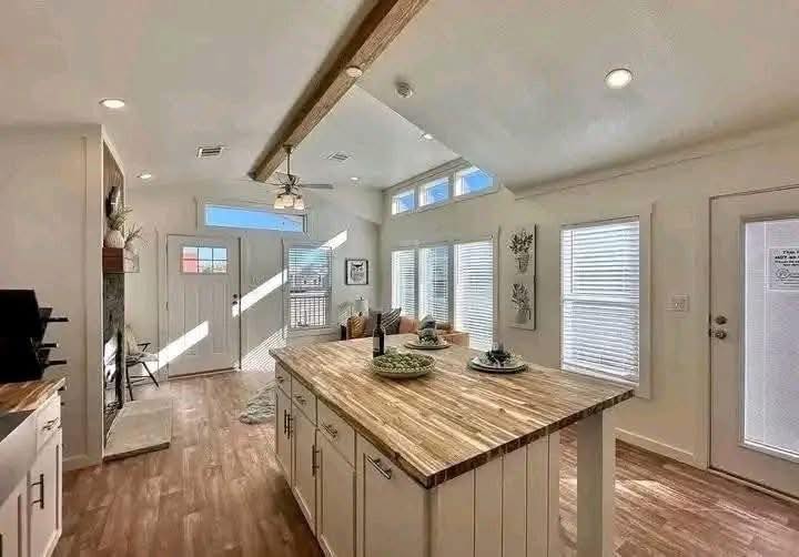 Spacious modern kitchen and living area with wooden floors, a large island, white cabinets, natural light from tall windows, and cozy decor.