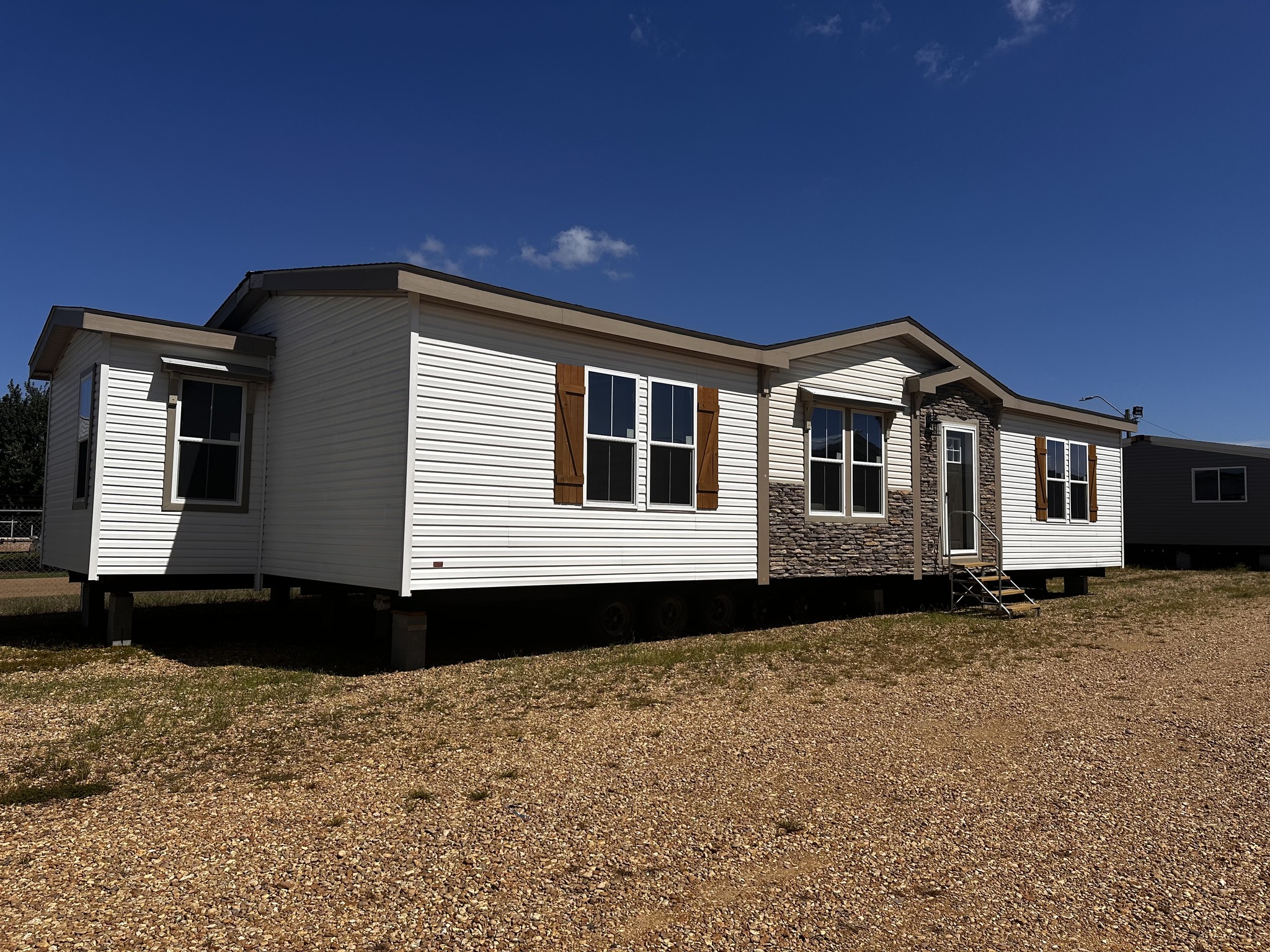 A white manufactured home with brown shutters and stone accents sits on a gravel lot under a clear blue sky, evoking a sense of rural tranquility.