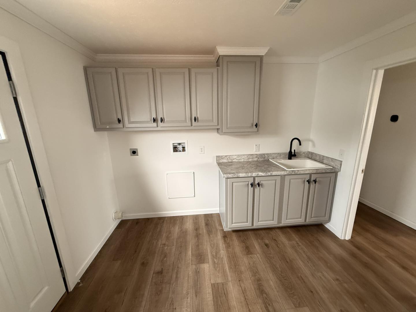 A clean, modern laundry room with light gray cabinets, white walls, and a wooden floor. It features a marble countertop, sink, and black faucet, conveying simplicity.