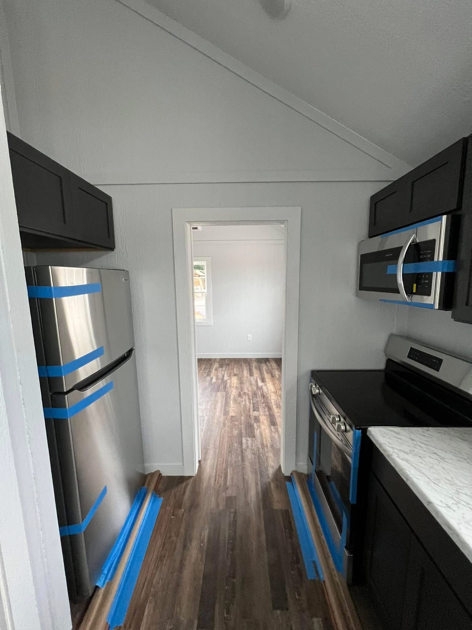 Compact, modern kitchen with dark cabinets, stainless steel fridge, and appliances marked with blue tape. The wood floor leads to a bright room.