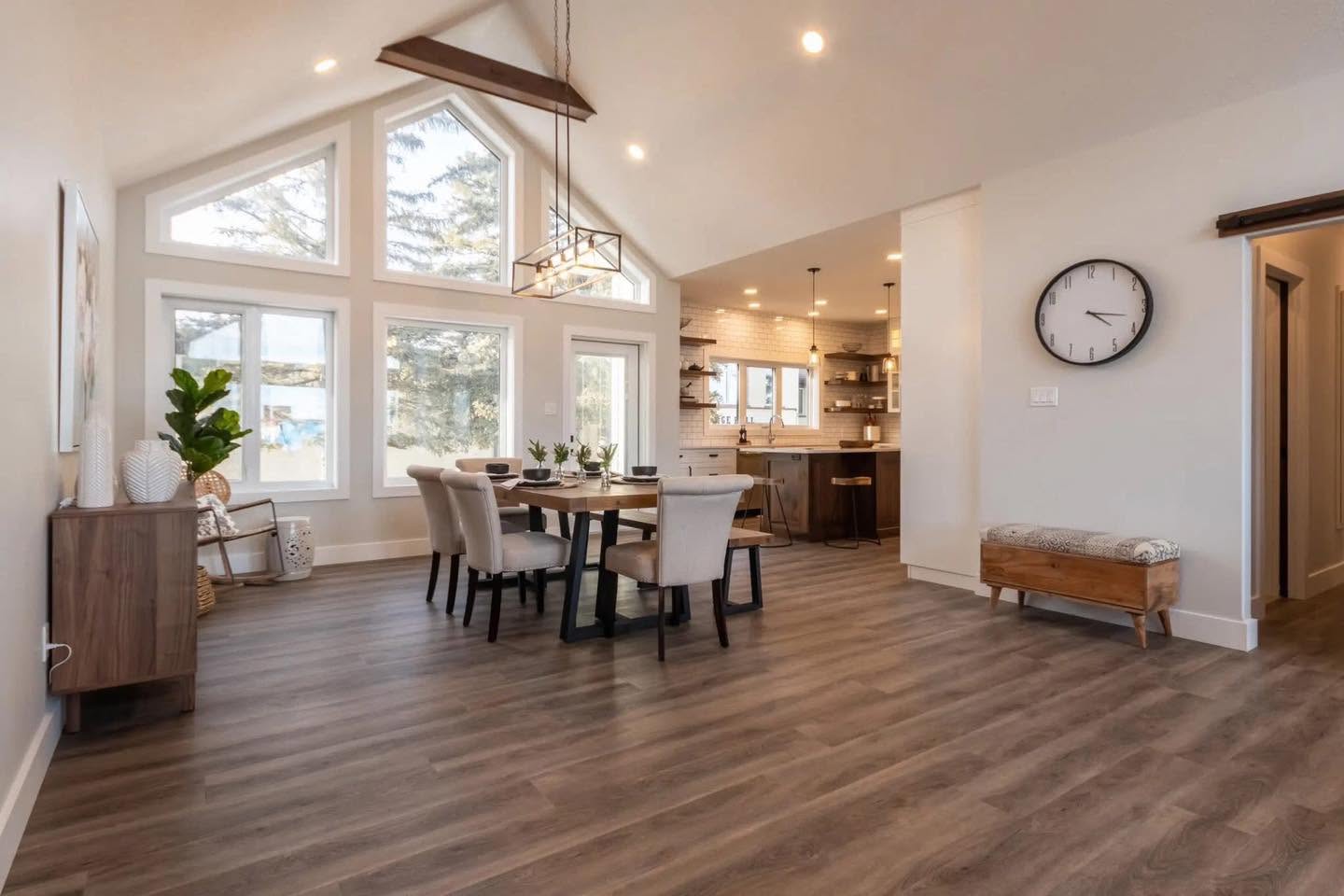 Spacious, modern dining area with high ceilings and large windows allowing natural light. Features a wooden table, white chairs, and a kitchen view.