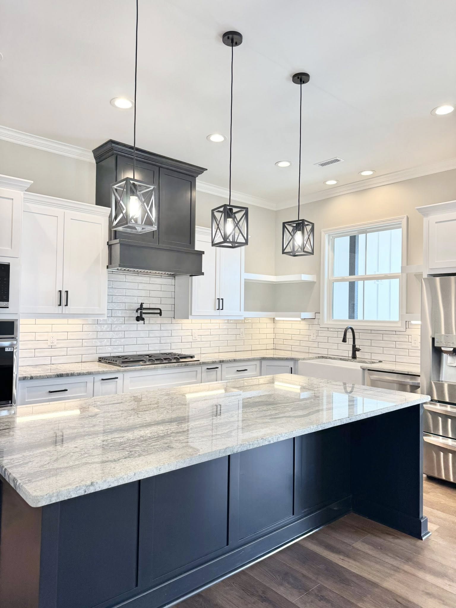 Spacious, modern kitchen with wooden floors, featuring a black island, white cabinets, built-in ovens, pendant lights, and a bright, airy feel.