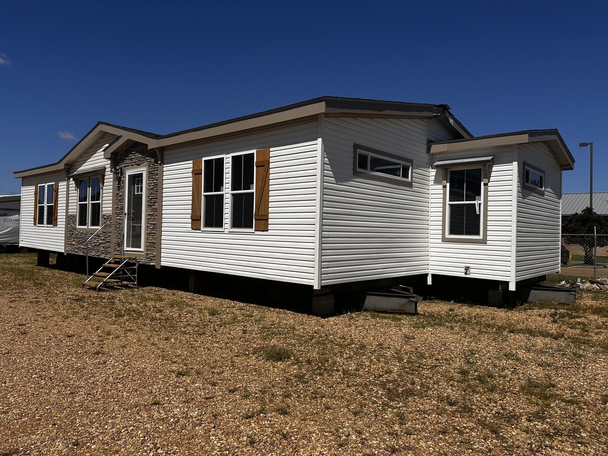 White modular home on a gravel lot, featuring multiple windows with wooden shutters, a stone accent near the front door, under a clear blue sky.