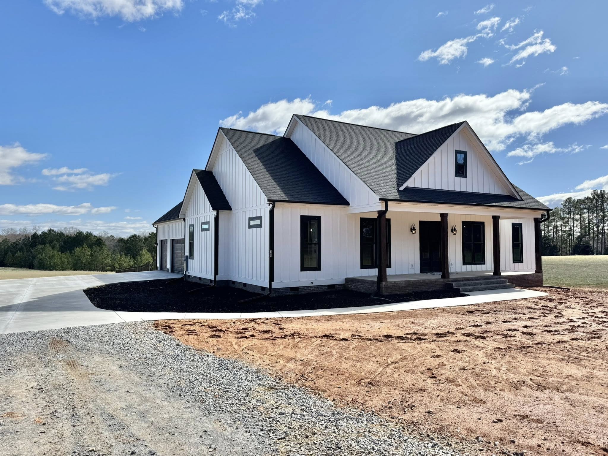 A modern farmhouse with white siding and a dark roof under a bright blue sky. The house features a front porch, large windows, and a garage, set in a rural landscape.