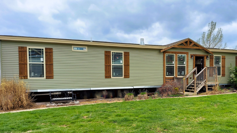 A beige manufactured home with wooden shutters sits on a lush green lawn under a cloudy sky. A small porch with stairs leads to the entrance, conveying a cozy, inviting feel.