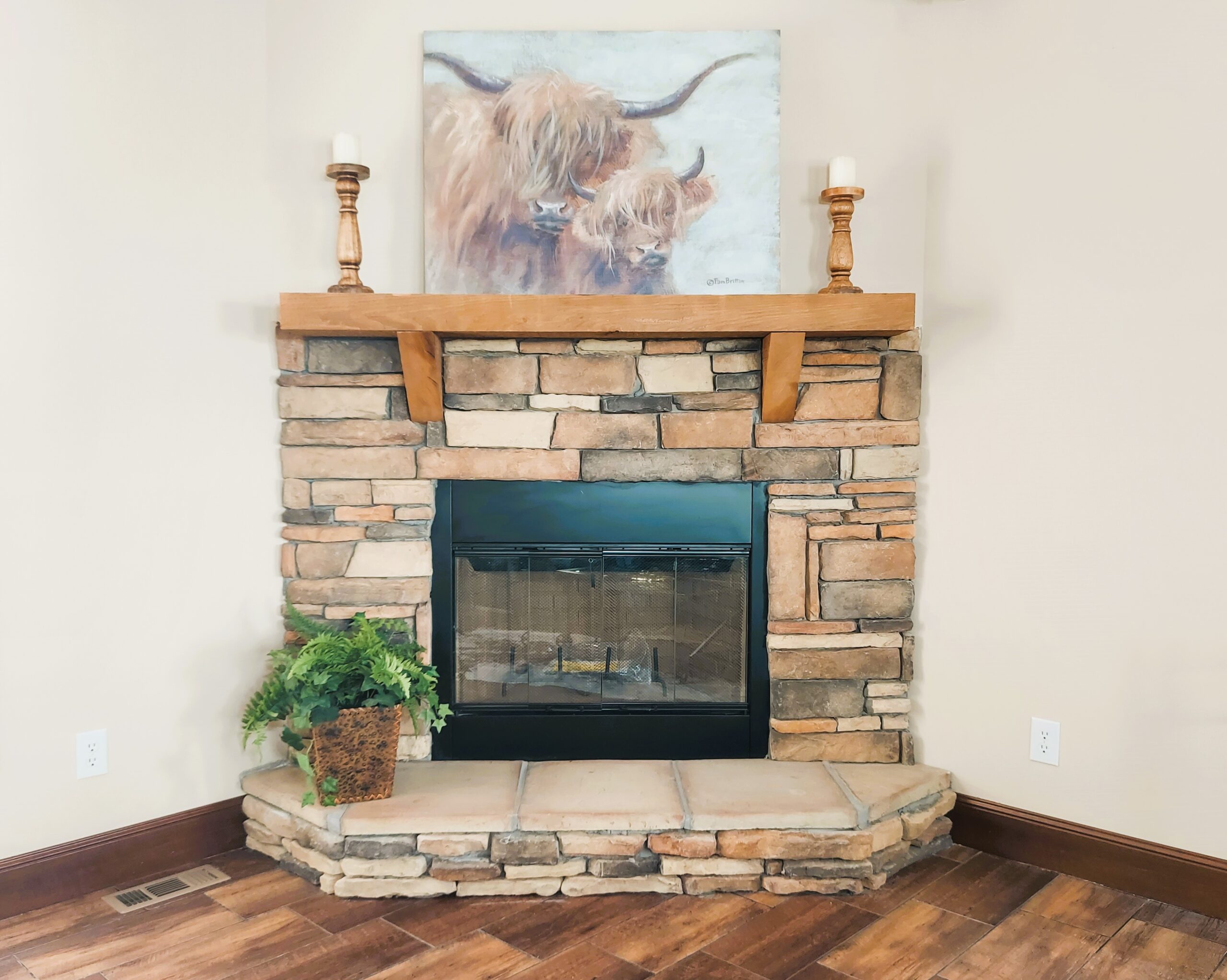 Cozy stone fireplace with wooden mantel, adorned by two candles and a painting of two shaggy Highland cows. A potted fern adds a natural touch.