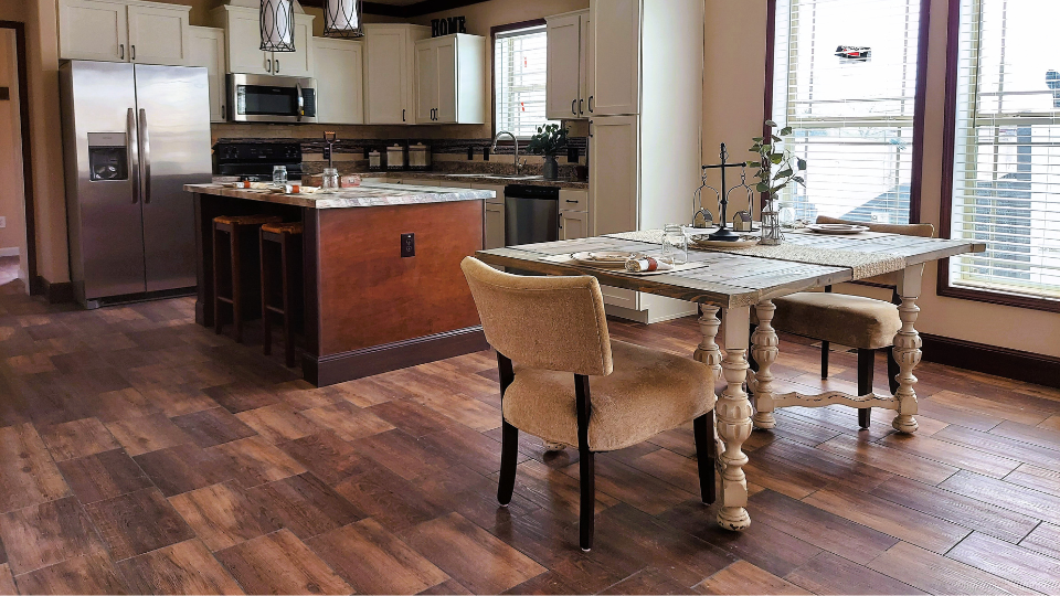 Bright kitchen with wooden flooring, featuring a white island with brown accents, stainless steel appliances, and a rustic dining table with padded chairs.