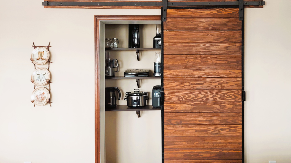 Pantry with a wooden sliding barn door partially open, revealing shelves with kitchen appliances. Three decorative plates hang on the left wall.