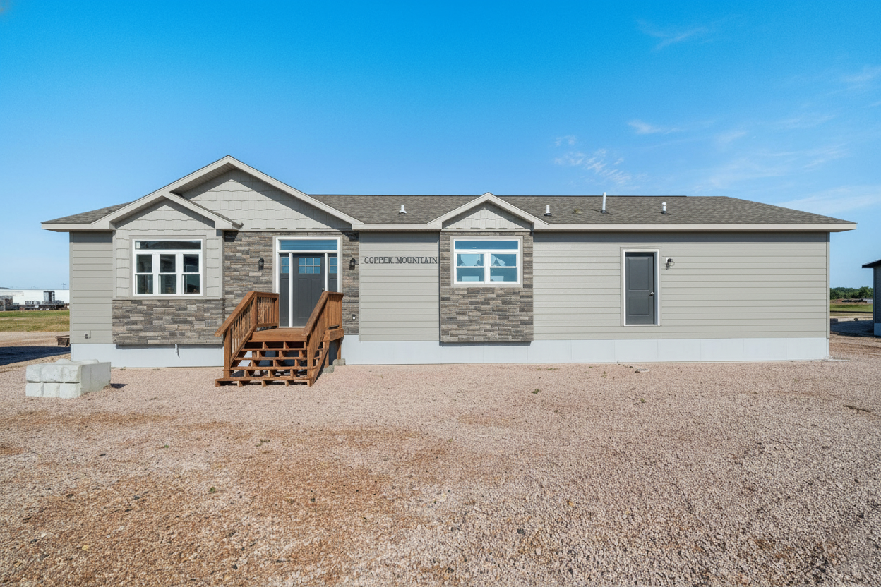 Single-story modular home with gray siding and stone accents, wooden steps leading to the door. It's set on a gravel lot under a clear blue sky.