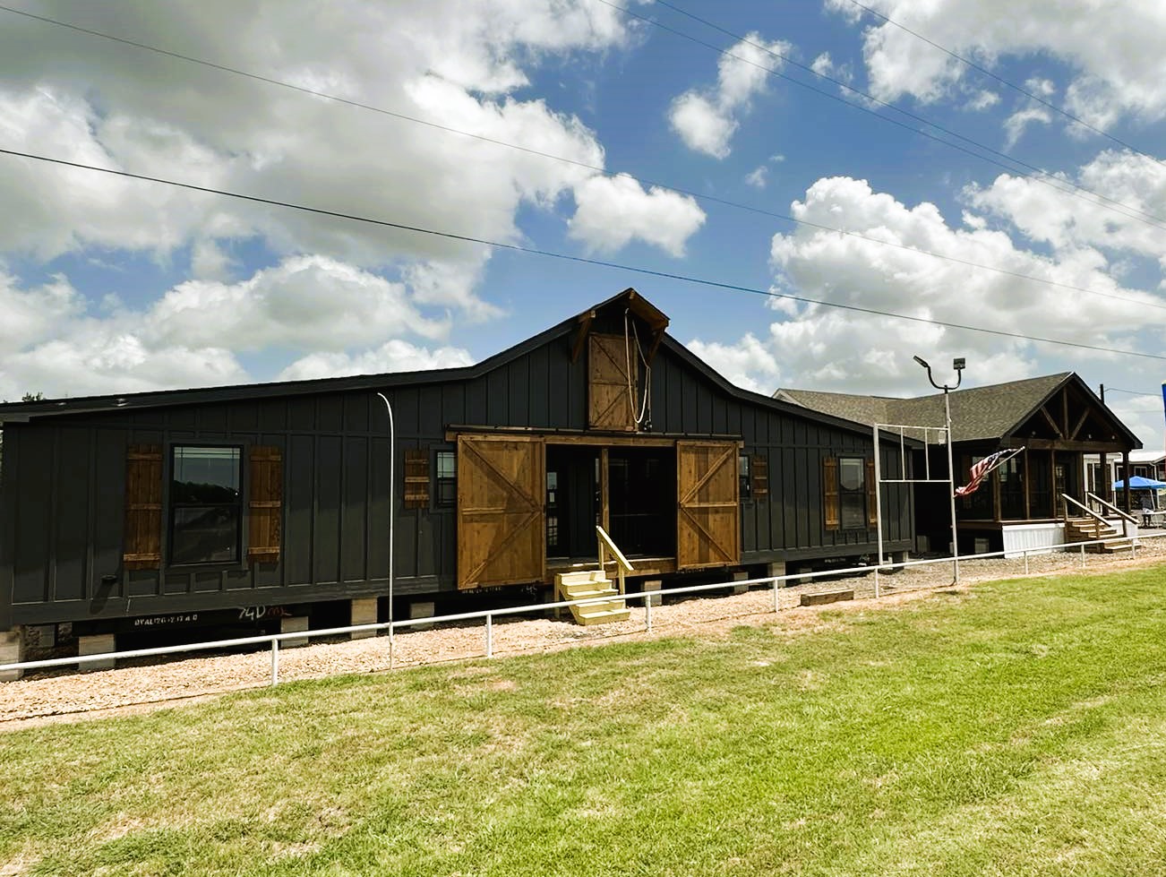 Long, dark gray barn-style building with wooden doors and windows under a partly cloudy sky. The vibrant green grass and path enhance a rural atmosphere.