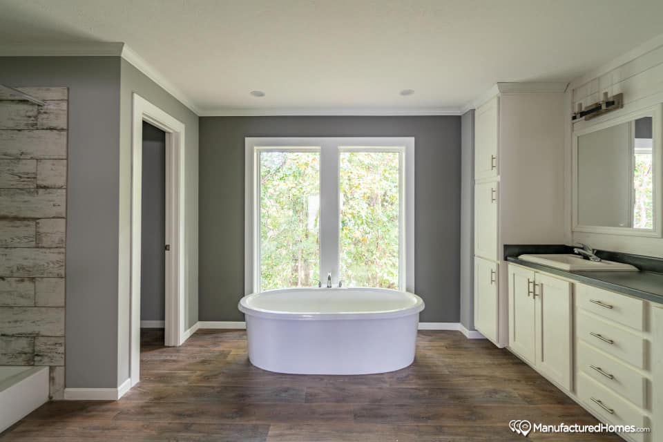 Spacious bathroom with a white oval bathtub centered below large windows. The room features gray walls, wood flooring, and white cabinetry, conveying a modern and serene atmosphere.