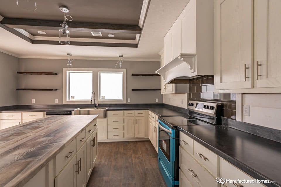 Modern kitchen with light wood cabinetry, dark countertops, and a blue oven. Two windows above a farmhouse sink and industrial light fixtures create a bright, inviting space.