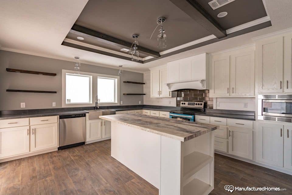 A modern kitchen with white cabinets, wood floors, and a central island. Stainless steel appliances are set against a gray accent wall with open shelves.